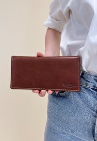 Osoba trzymająca brązowy skórzany portfel z widocznym przeszyciem i wytłoczonym napisem "Gusti Leder studio" w rogu, ubrana w białą koszulę i niebieskie dżinsy.