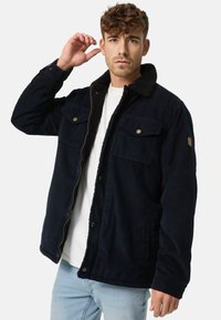 Marineblauwe corduroy jas met een sherpa kraag, voorzien van twee borstzakken en knopenaccenten, gecombineerd met een wit t-shirt en lichtblauwe jeans.