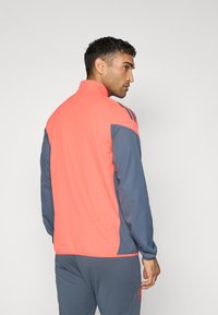 Sportjacket met een koraalkleurig bovenstuk en marineblauwe mouwen. Gemaakt van lichtgewicht materiaal met een hoge kraag en drie strepen op de schouders.