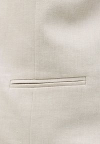 Gros plan d’un tissu beige avec une poche horizontale fendue et des coutures visibles sur un matériau texturé.