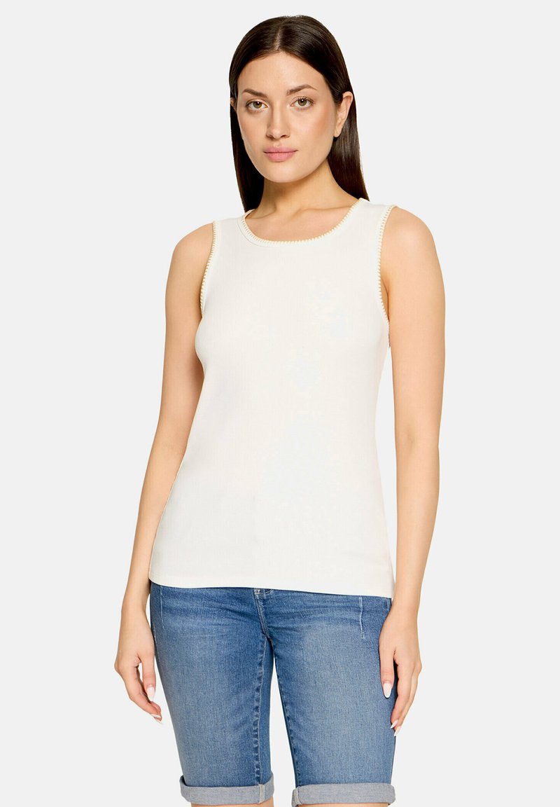 Femme aux cheveux raides et foncés portant un haut blanc sans manches et un jean bleu retroussé, debout devant un fond clair.