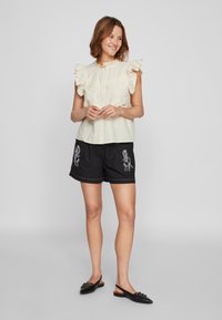 Kremfarget bluse med korte ermer, rysjer og blondekant, kombinert med svarte shorts med hvite broderte mønstre. Svarte slip-on sko.