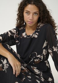 Melnās ziedu kleitas ar garām caurspīdīgām piedurknēm, ar pieguļošu jostu un pleitu dizainu. Ziedu raksti ir redzami bēša un brūnā toņos.