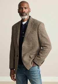 Brown houndstooth blazer in textured fabric, single-button closure, worn over a dark, collared shirt, paired with light blue jeans.
