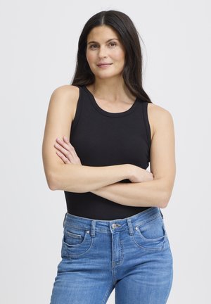 Femme aux longs cheveux noirs portant un haut noir sans manches et un jean bleu, debout les bras croisés devant un fond uni.