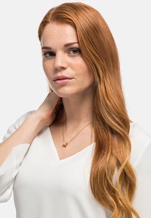 Collier en or avec un pendentif rond orné d'une pierre précieuse claire, porté sur une blouse blanche à col en V, cheveux longs et ondulés, de couleur rouge.