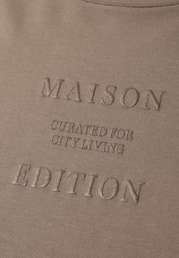 Texte en relief "MAISON EDITION" avec un plus petit "CURATED FOR CITY LIVING" sur un tissu beige, montrant la texture et les coutures près de l'encolure.