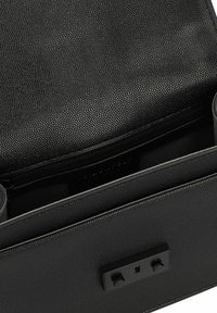 Bolso de cuero negro texturizado con una forma estructurada, que cuenta con un cierre de cremallera y un acento de hardware negro plano en la parte frontal.