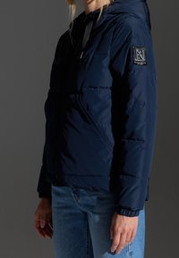 Marineblå pufferjakke med hætte, quiltet tekstur, lynlås foran og et logo patch på venstre ærme. Båret over denim jeans.