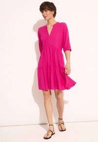 Różowa sukienka do kolan z dekoltem w kształcie litery V i rękawami o długości trzech czwarci. Posiada warstwową spódnicę z falbanami oraz pojedynczy guzik poniżej dekoltu.
