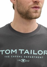 Gray cotton t-shirt featuring "TOM TAILOR" logo in light blue. Crew neck with a smooth texture. Short sleeves, close-fitting design.