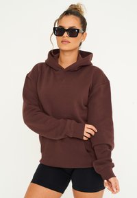 Braun gefütterter Kapuzenpullover mit lockerer Passform, langen Ärmeln, gerippten Bündchen und einer Fronttasche, kombiniert mit schwarzen Shorts und Sonnenbrille.