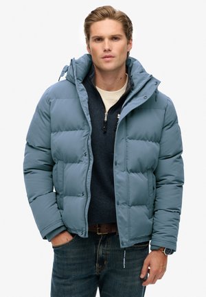 Lysblå pusteblækjakke med hætte, der har polstrede sektioner, en frontlynlås og trykknaplukninger, båret over en marineblå sweater.