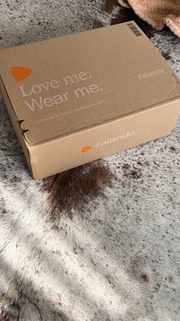 Zalando kartong märkt "Älska mig. Bär mig." i vit text med en orange logotyp, som vilar på en strukturerad, ljus yta.