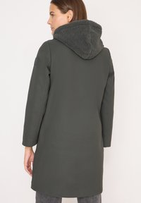 Cappotto verde scuro con cappuccio foderato in pelliccia e texturizzato. Presenta maniche lunghe e arriva a metà coscia. Design semplice e moderno con accenti minimi.
