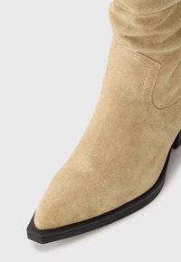 Bottine en daim beige avec un bout pointu, un design froncé et une semelle en caoutchouc noir, présentant de subtils détails de couture sur la partie supérieure.