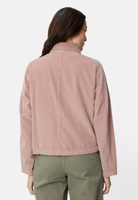 Kurta kurtka z różowego sztruksu, z kołnierzem, prążkowaną fakturą i prostym krojem, zestawiona z oliwkowozielonymi spodniami. Podkreślony widok od tyłu.