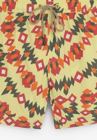 Un maillot de bain jaune avec un motif abstrait en zigzag et en diamant en rouge, orange et gris. Il est doté d'un cordon de serrage et d'une texture de tissu douce.
