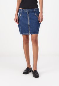 Denim miniskjørt med en tettsittende fasong, utstyrt med glidelås foran, to side lommer med glidelås, og en mørkeblå vask. Kombinert med svarte loafer-sko.