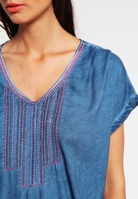 Niebieska bawełniana tunika z dekoltem w kształcie litery V, ozdobiona haftem w czerwonych i czarnych nitkach wzdłuż przodu. Krótkie rękawy.