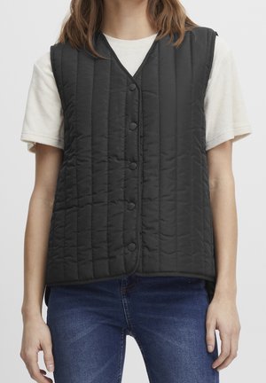 Woman wearing a black quilted buttoned vest over a white short-sleeve shirt and blue jeans, standing against a plain background.