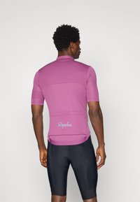 Kurze Ärmel Radtrikot aus sanftem rosa Stoff mit hohem Kragen, Rückentasche und Logodetail in hellblau, kombiniert mit schwarzen Shorts.