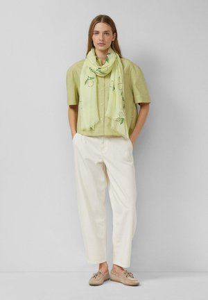 Donna in piedi contro un muro grigio che indossa una camicia verde chiaro a maniche corte, pantaloni crema, scarpe beige e una sciarpa gialla con motivi di limoni.