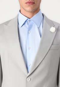 Lichtgrijze colbert met notch-lapels, gecombineerd met een lichtblauw overhemd. Kenmerkt zich door een witte bloemenlaping en twee knopen.