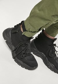 Zapatillas altas negras con parte superior de tela texturizada, punta redonda, suela gruesa y cordones, combinadas con pantalones verdes con puños.