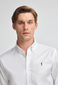 Vit skjorta med knappar som har en klassisk krage, texturerad tyg och en liten broderad logotyp på vänster bröst.