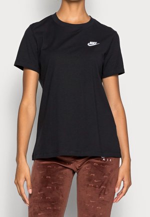 Personne portant un t-shirt Nike noir et un pantalon Nike Air marron avec des logos répétés, debout devant un fond clair uni.