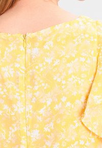 Bakvy av en person som bär en gulblommig klänning med en centrerad dragkedja bak och en något av axeln nacke.