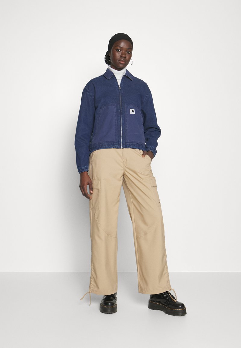 Giacca di denim blu con zip e due tasche frontali, indossata sopra un dolcevita bianco e pantaloni cargo beige, abbinata a stivali neri.