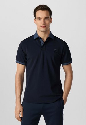 Jonge man die een donker marineblauw polo met korte mouwen draagt met een contrasterende kraag en mouwranden, staand tegen een effen lichte achtergrond.