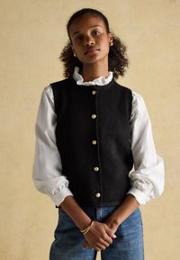 Woman with curly hair wears a white blouse with ruffled collar and cuffs, a black vest with gold buttons, blue jeans, and gold jewelry.