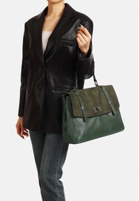 Borsa a tracolla verde in pelle con pattina in camoscio, hardware in argento e doppi manici, posizionata su un blazer nero e jeans blu.