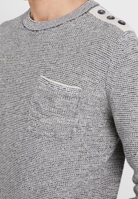 Grauer Strickpullover mit runder Halslinie, strukturiertem Muster, einer Brusttasche und drei großen Knöpfen an der Schulter.