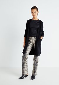 Svart cardigan med öppen front och fickor, bärs över en mörk topp och mönstrade beige byxor med svarta tigerränder. Svarta klackskor.