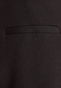 Gros plan d'une veste en tissu noir avec un détail de poche passepoilée horizontale centrée dans le cadre.