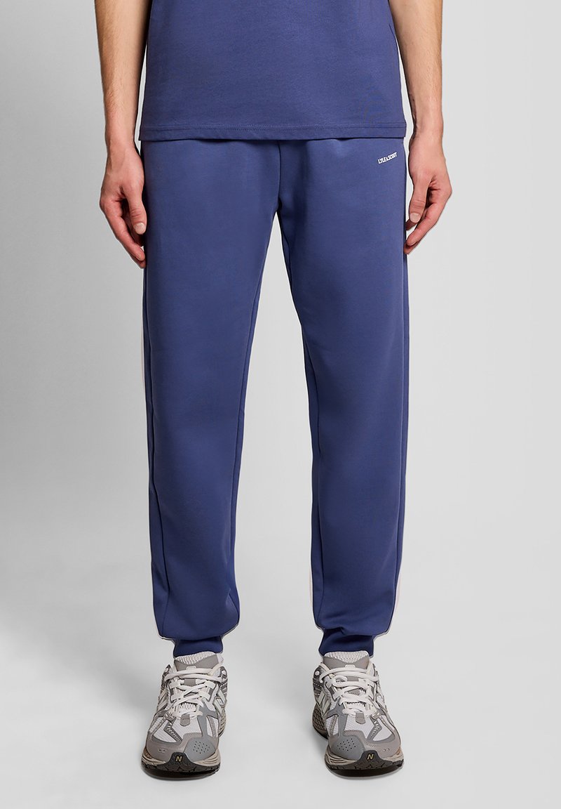 Personne portant un pantalon de survêtement bleu et des baskets gris-blanc, debout contre un fond uni gris clair.