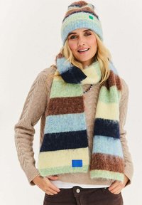 Zestaw dzianinowej wełnianej czapki i pasującego szalika w pastelowe paski w kolorach niebieskim, zielonym, brązowym i granatowym, z małą niebieską metką na szaliku.