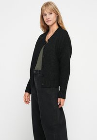 Svart strikket cardigan med en teksturert chevron-mønster, med rund hals, knappelukking og ribbestrikkede mansjetter. Brukt over en grønn topp.