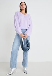 Camisola lilás de malha com decote em V, mangas compridas e corte descontraído, combinada com jeans azul claro e saltos prateados de bico fino, segurando um lenço de ganga.