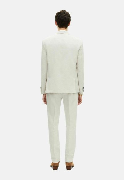 Completo beige chiaro, con una vestibilità su misura e una texture liscia. Dettagli notevoli includono una fessura singola e bottoni scuri sulle maniche. Scarpe marroni.