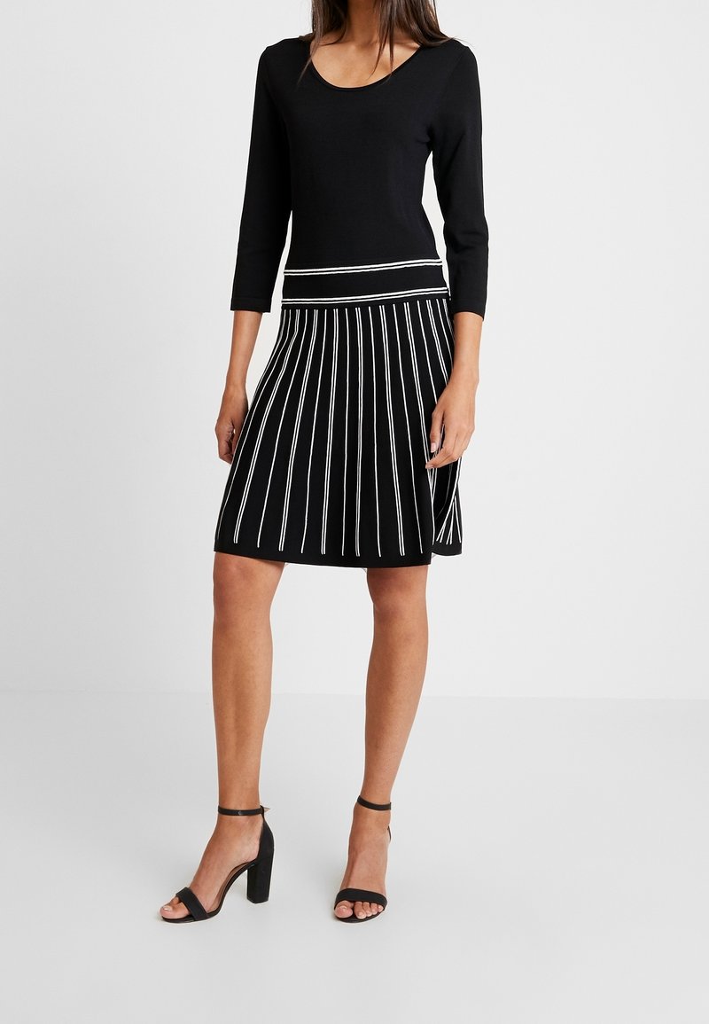 Femme portant une robe noire à manches trois-quarts et une jupe plissée avec des rayures blanches verticales, associée à des sandales noires à talons hauts.