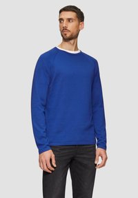 Blauer langärmeliger Pullover mit Rundhalsausschnitt, aus strukturiertem Stoff gefertigt. Mit gerippten Bündchen und Saum, kombiniert mit dunklen Jeans.