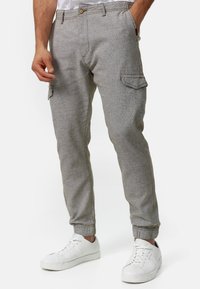 Man in lichtgrijze cargobroek met knoop- en klepzakken, gecombineerd met witte sneakers op een witte achtergrond.