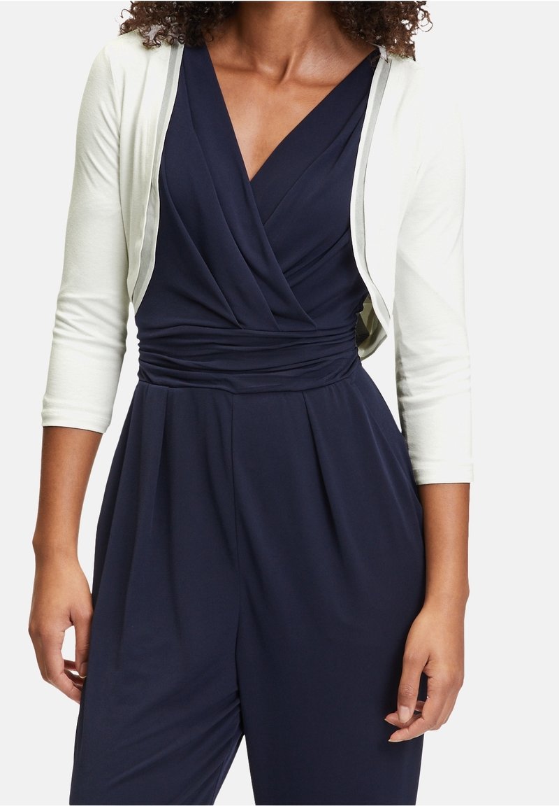 Femme portant une robe portefeuille bleu marine avec une taille froncée et un cardigan court blanc à manches trois-quarts.