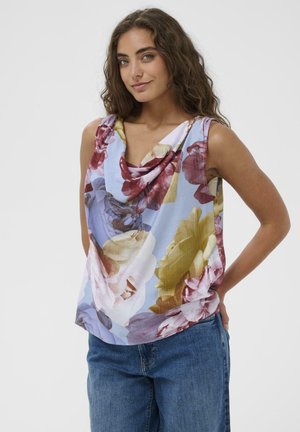 Kobieta o falowanych włosach, ubrana w bezrękawnik z kwiatowym nadrukiem i dekoltem z marszczeniem oraz niebieskie dżinsy, stojąca na jednolitym tle.