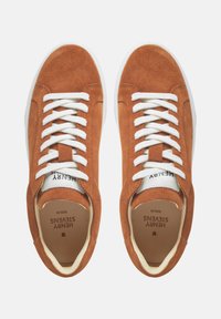 Sneakers in suede marrone con lacci bianchi, un interno crema e una suola in gomma bianca. Presentano un'etichetta del brand sulla linguetta e sulla soletta.